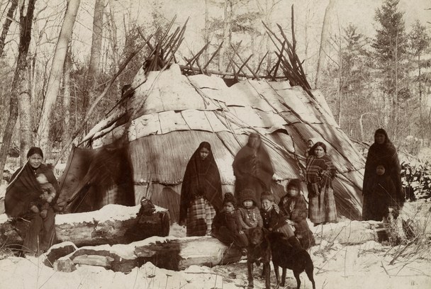 Women and children in blankets in front of a large domed bark lodge in snowy woods on the St. Croix, about 1885
