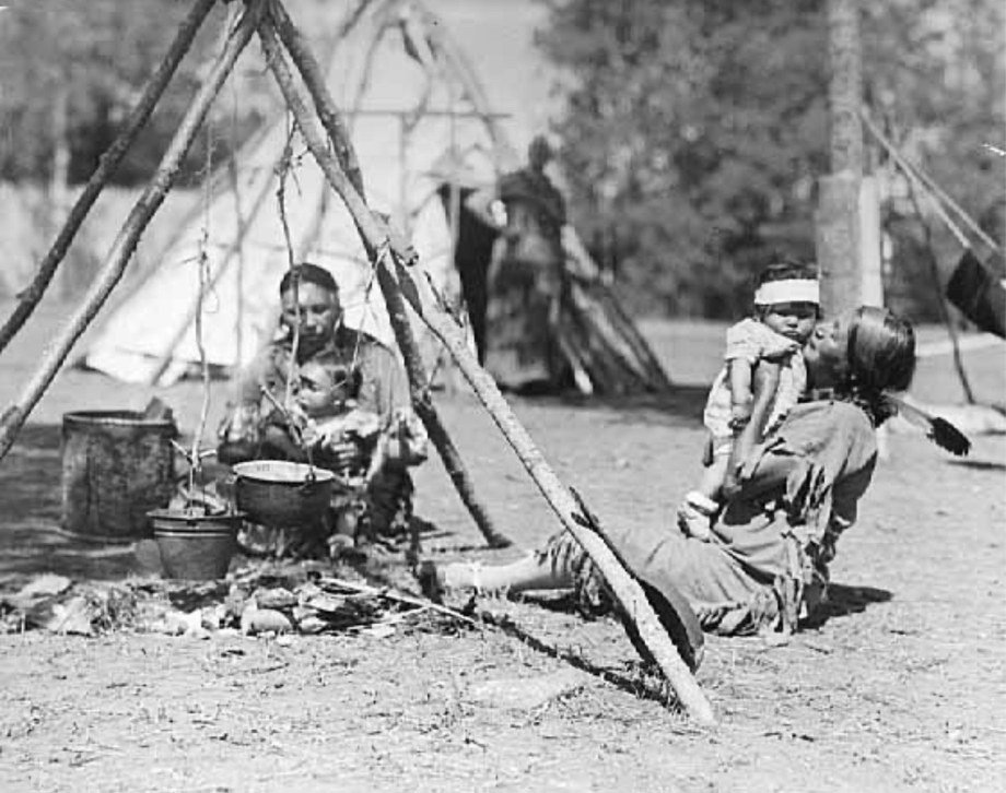 Women and children seated around a camp fire with a kettle on a tripod, Chippewa village, Itasca State Park, July 1926