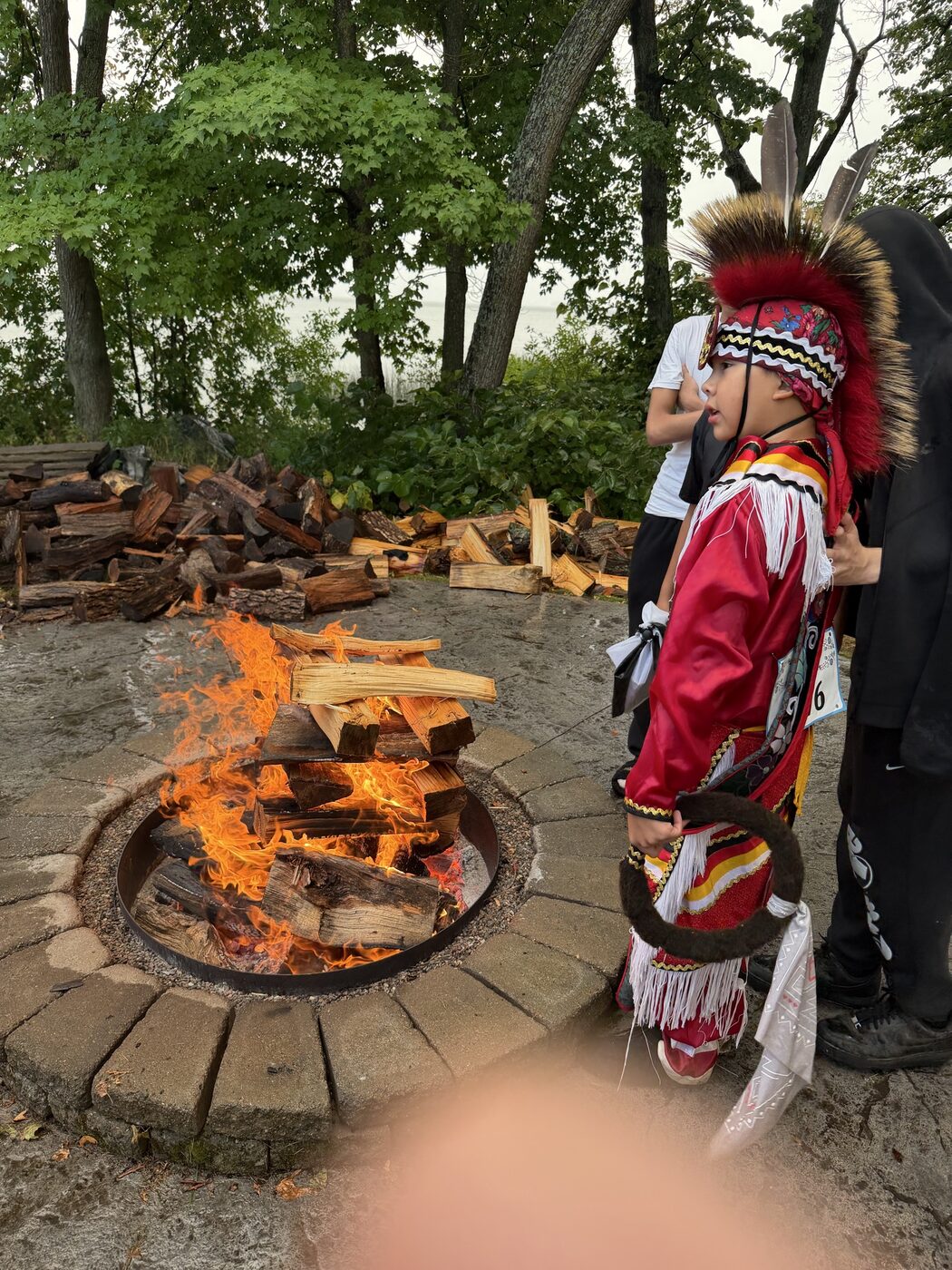 Young dancer in red regalia standing beside the fire